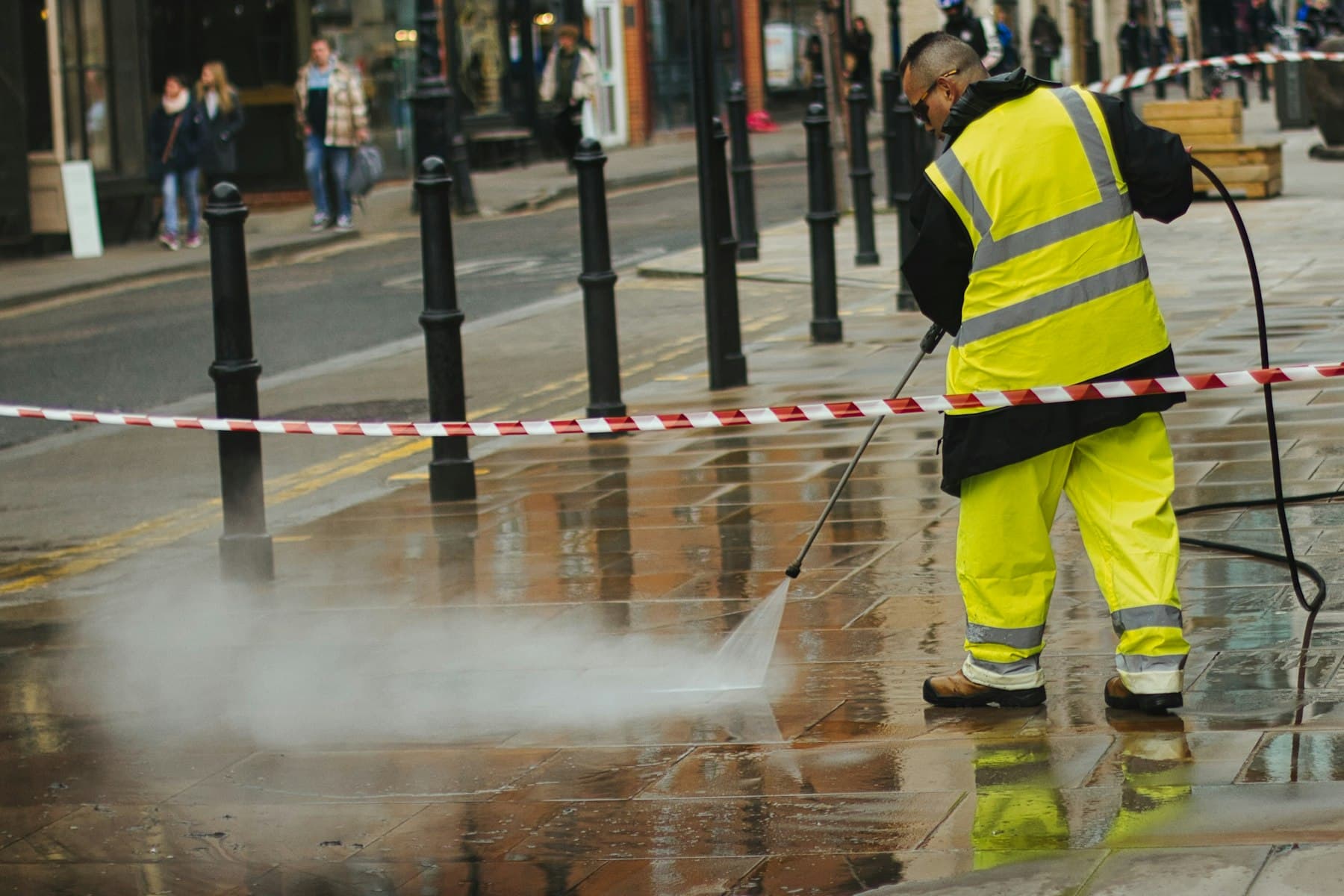 A worker in a high-visibility suit pressure washing a paved street.